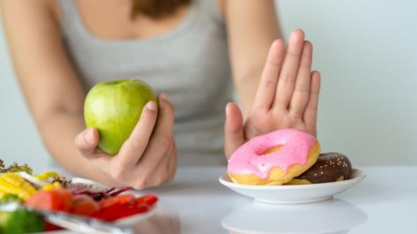A woman holding an apple while saying "no" to a plate of donuts